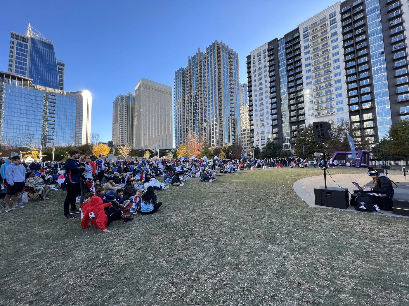 Spectators seated on a park lawn in Uptown Charlotte at dusk with a stage and high-rises behind