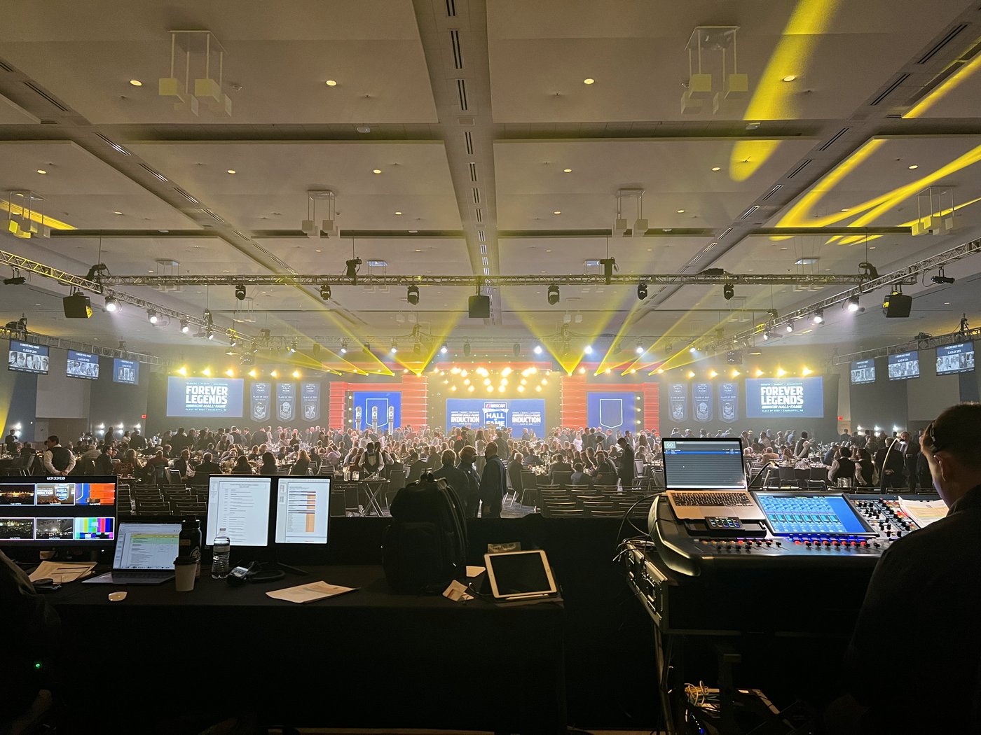 The induction ballroom from behind the show control desk, with video and audio consoles in the foreground and yellow beams over the stage