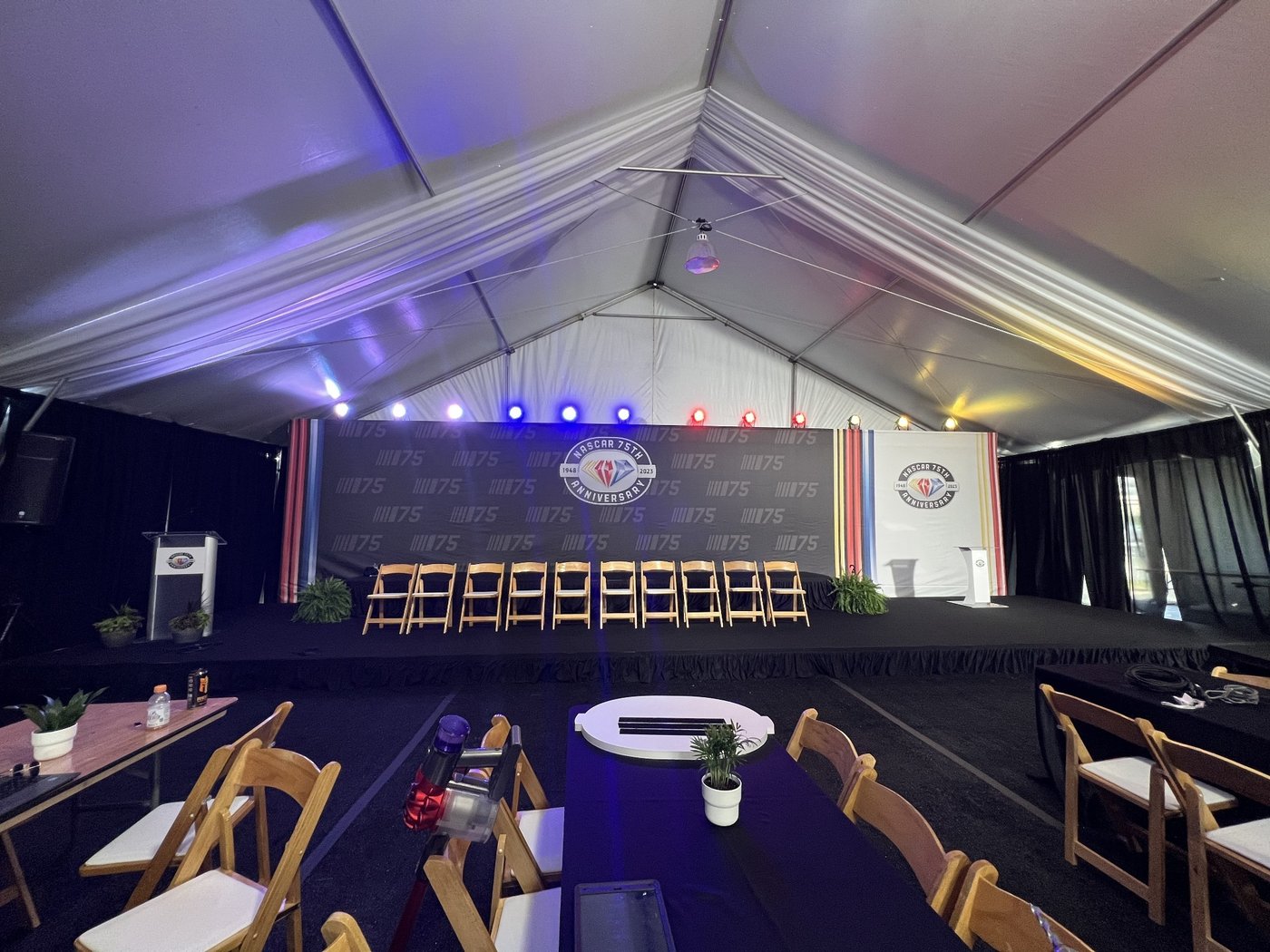Hospitality tent interior with a stage backdrop of NASCAR 75 graphics, a podium, rows of wood chairs and long tables