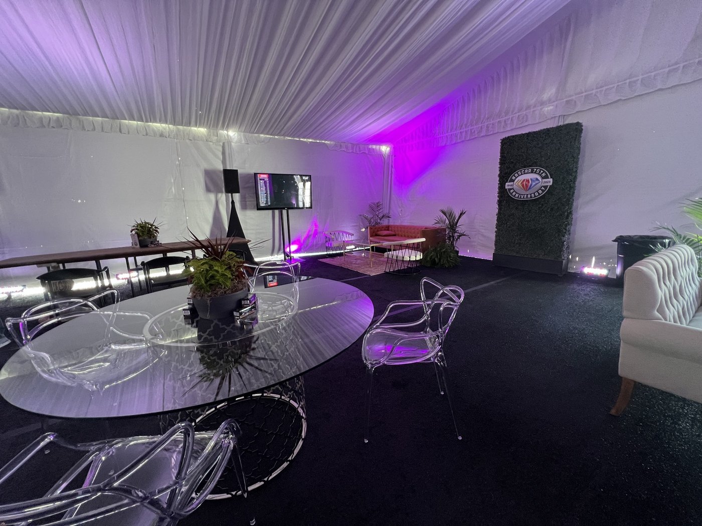 Tent lounge lit in purple with acrylic chairs, a glass table, a green wall logo and a television