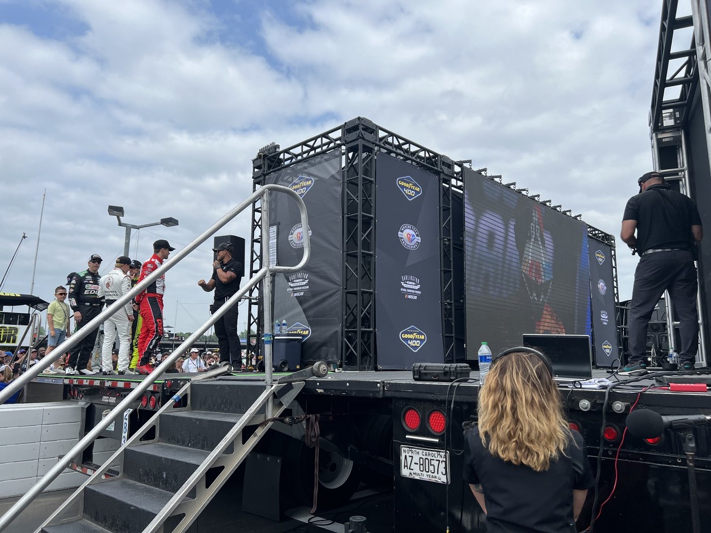 Drivers on a hauler stage at Darlington under a Goodyear 400 truss and LED screen, with Alli Kaser in a headset in the foreground