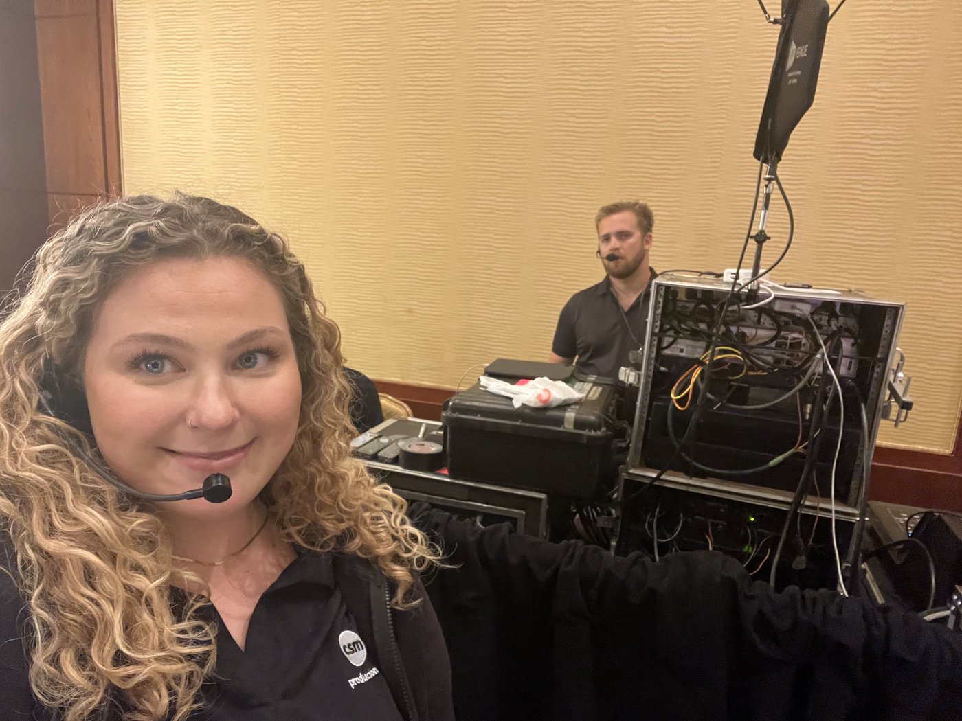 Alli Kaser smiling in a headset in front of racked video equipment, with a colleague wearing a headset behind