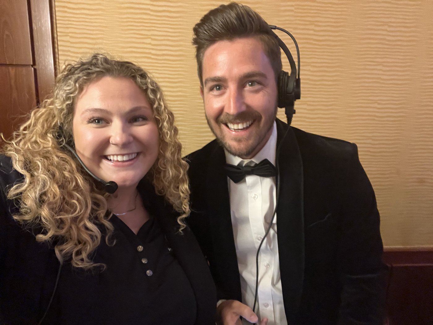 Alli Kaser and a colleague in a tuxedo, both wearing headsets, smiling for a selfie