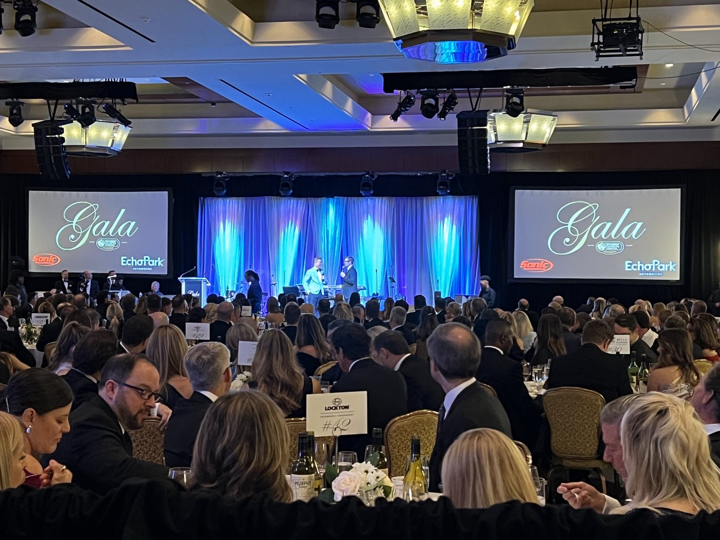 Gala ballroom with a lit blue-draped stage, two large screens and guests seated at tables