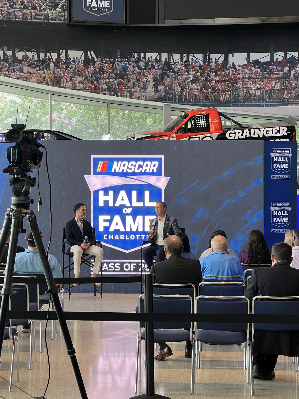 Two people in conversation on stage in front of a NASCAR Hall of Fame Class of 2024 backdrop, with a camera in the foreground