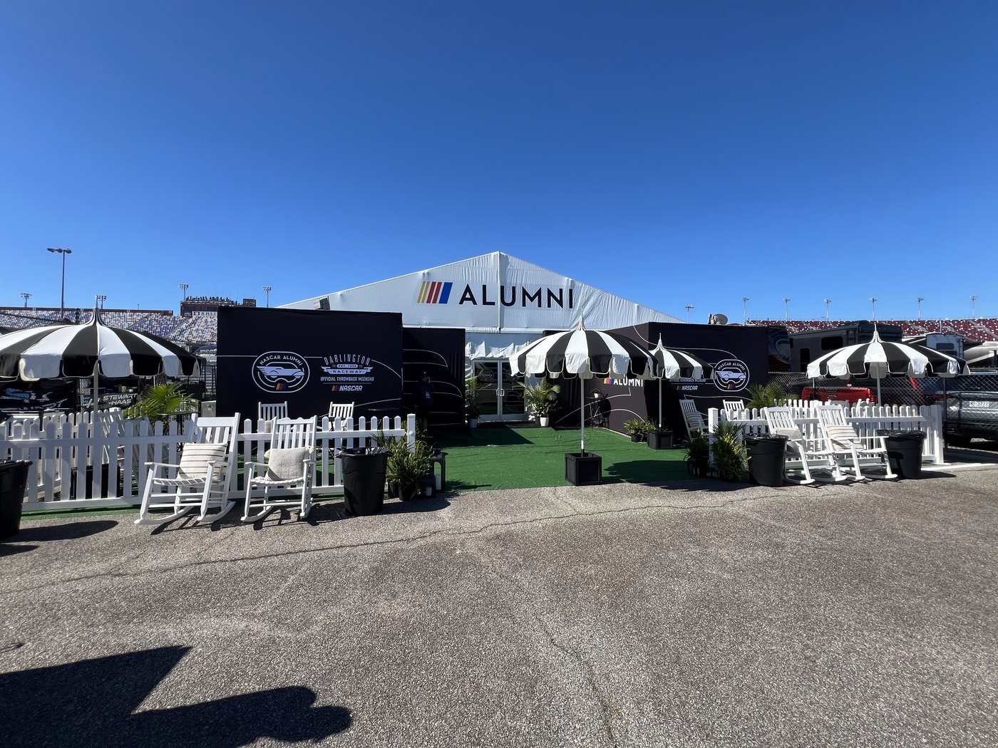 NASCAR Alumni hospitality tent at Darlington Raceway with striped umbrellas, rocking chairs and a white picket fence