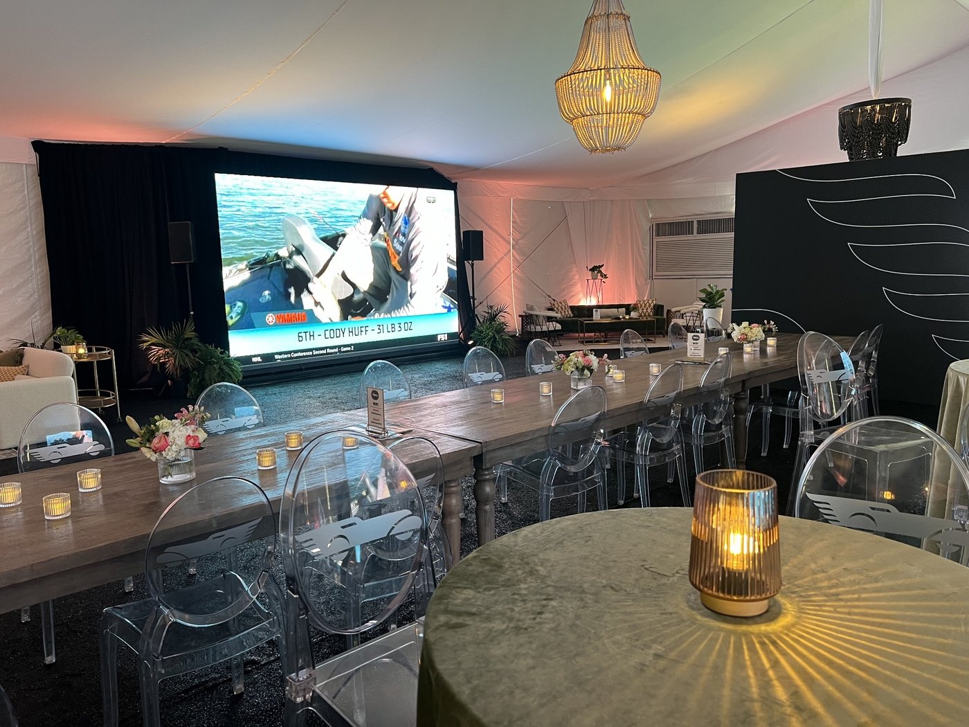 Long wood banquet table with clear chairs, candles and a chandelier, facing a large screen showing a boat race