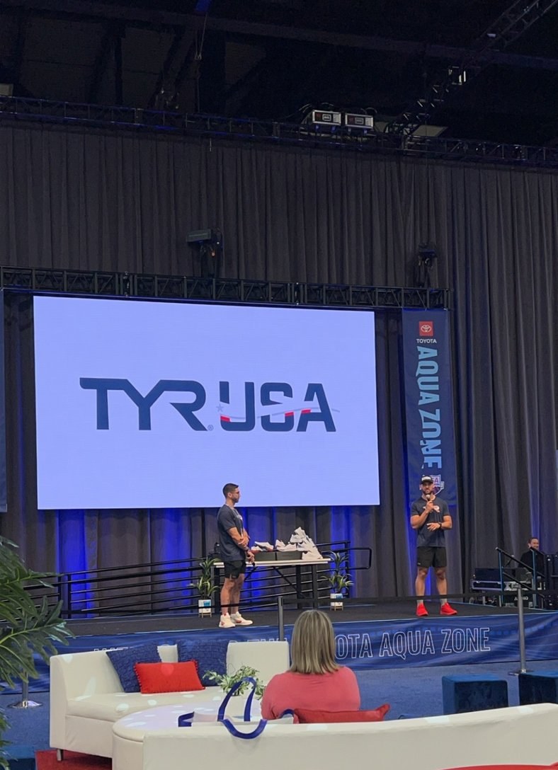A TYR USA screen above a small stage and a red-and-white lounge area at the Olympic Trials