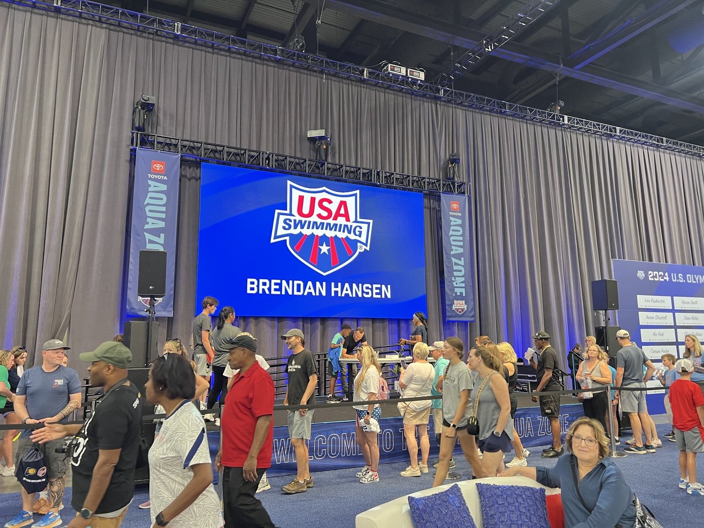The Aqua Zone stage screen showing a USA Swimming logo and Brendan Hansen's name as fans line up in front