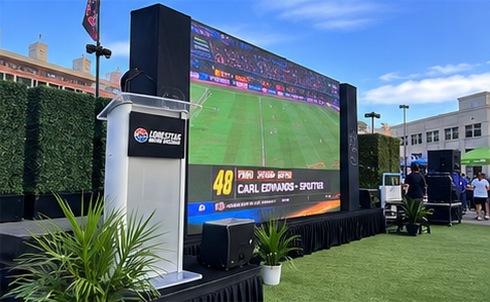 A podium in front of a large LED screen showing a live broadcast, on artificial turf with plants