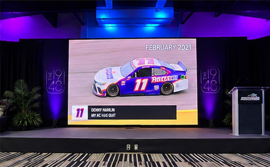 A stage with a large LED screen showing a race car graphic between two The 1948 panels, lit in purple and blue