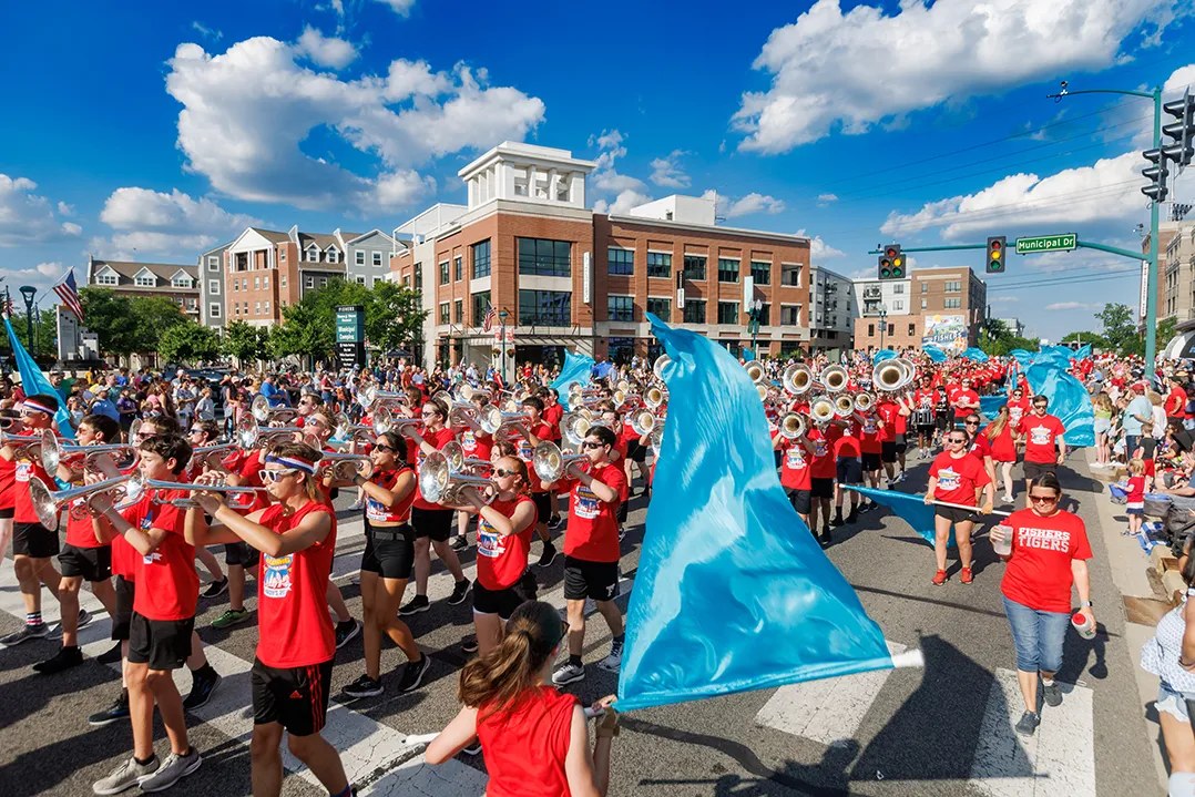 Marching band members in red shirts and a blue flag line moving down a Fishers street during the Spark! Fishers parade, with crowds along the curb