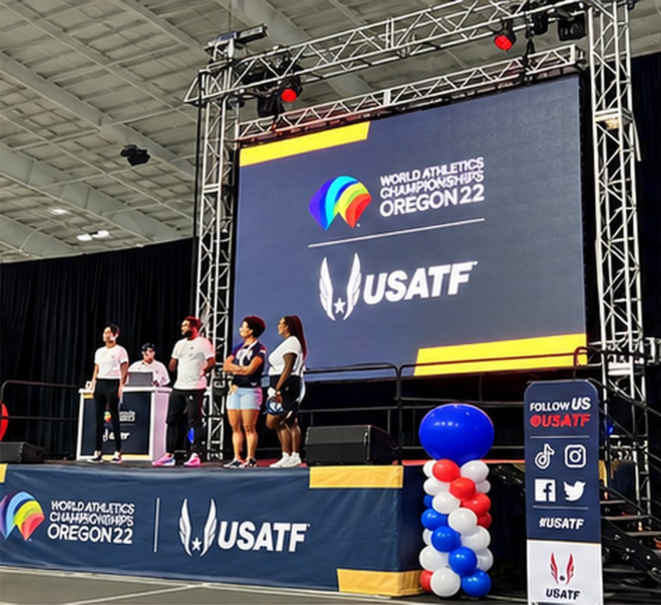 Four people standing on the USATF stage in front of a large screen, with a social media banner and red, white and blue balloon columns