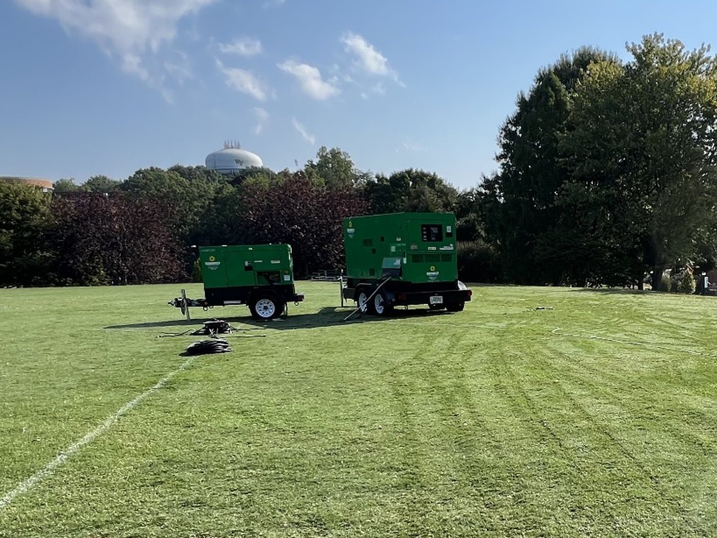Two green trailer-mounted generators set up on a lawn on a sunny morning