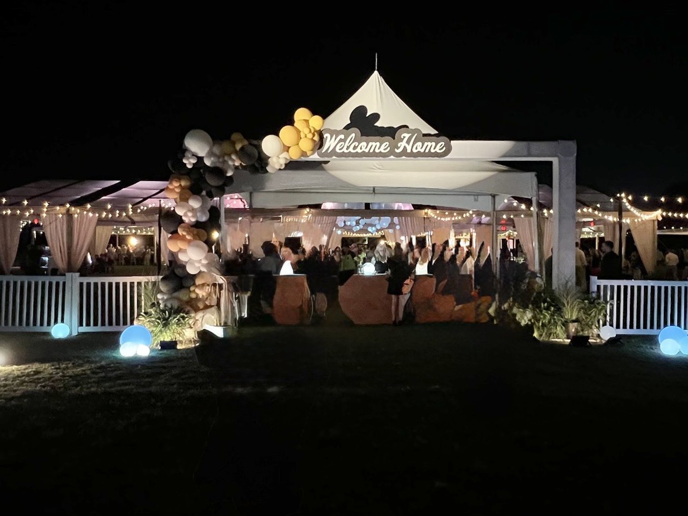 An outdoor tent lit with string lights at night, with a Welcome Home sign and balloon arch at the entrance